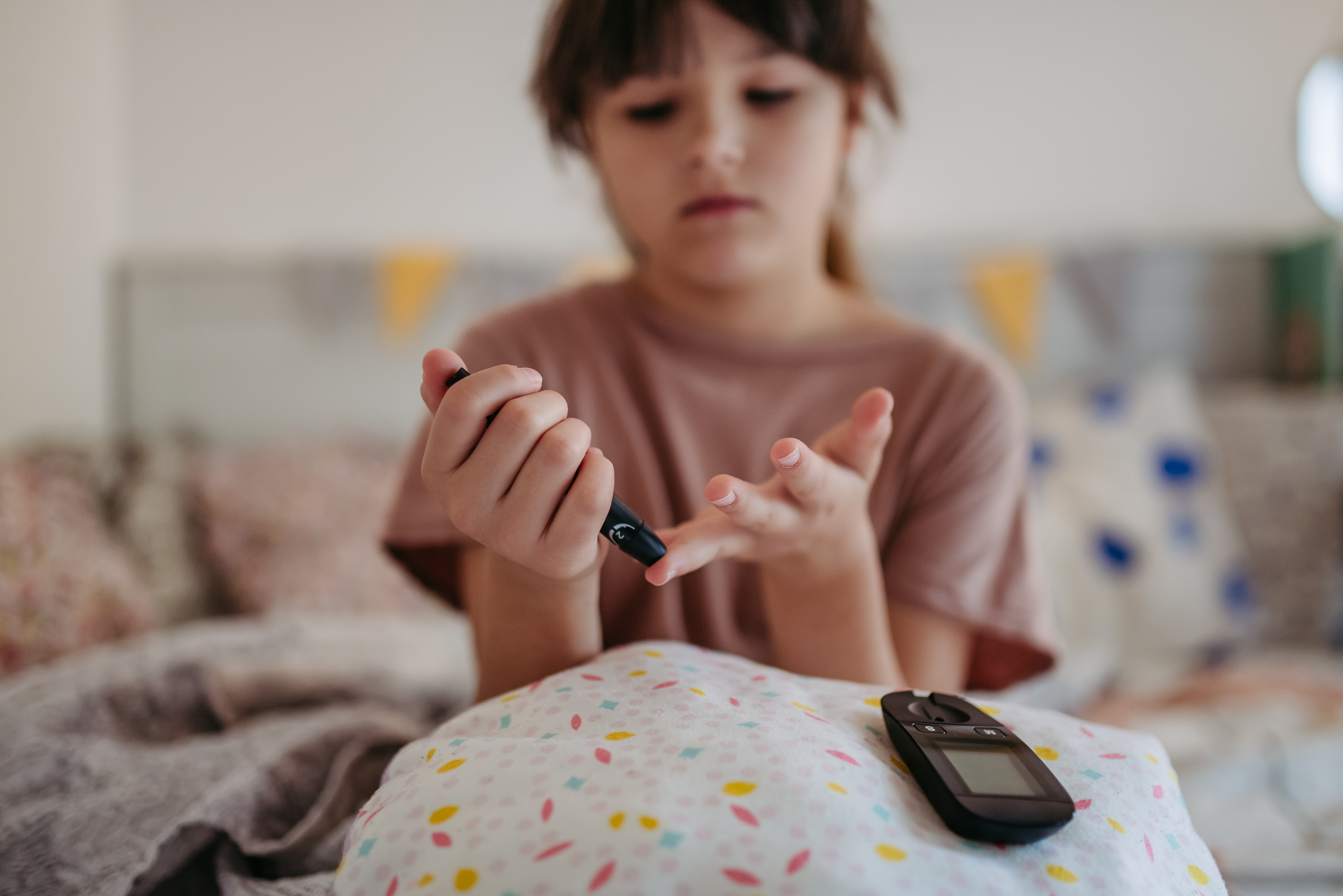 A imagem mostra uma pessoa sentada em uma cama, segurando um medidor de glicose em uma das mãos e fazendo um furo no dedo da outra mão. A imagem retrata um processo comum usado por pessoas com diabetes para monitorar seus níveis de açúcar no sangue. 